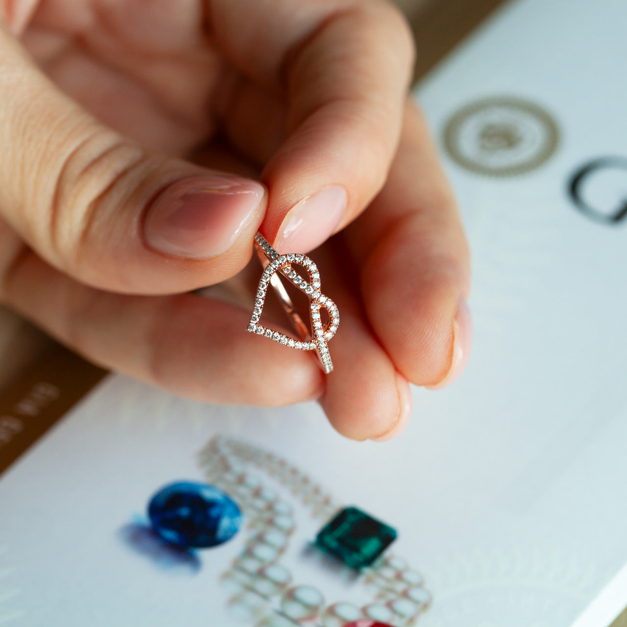 Hand holding a sparkling 18k white and rose gold heart-shaped diamond ring from a 2-piece ring set above a jewelry catalog with images of blue and green gemstones.